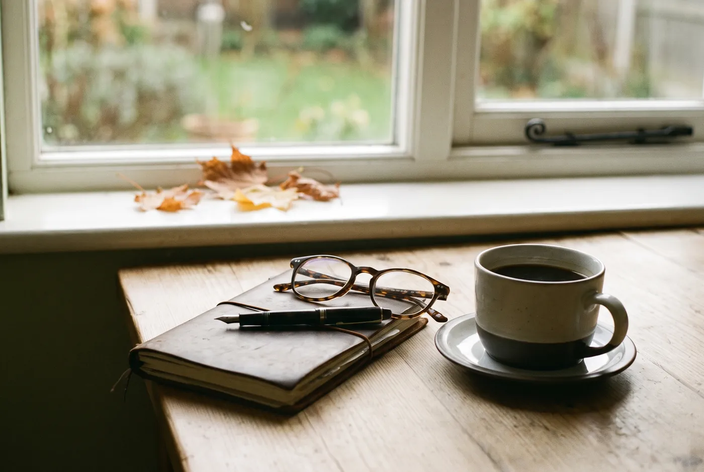 A closed notebook, a pen, reading glasses and a cup of coffee on a kitchen table in soft daylight.