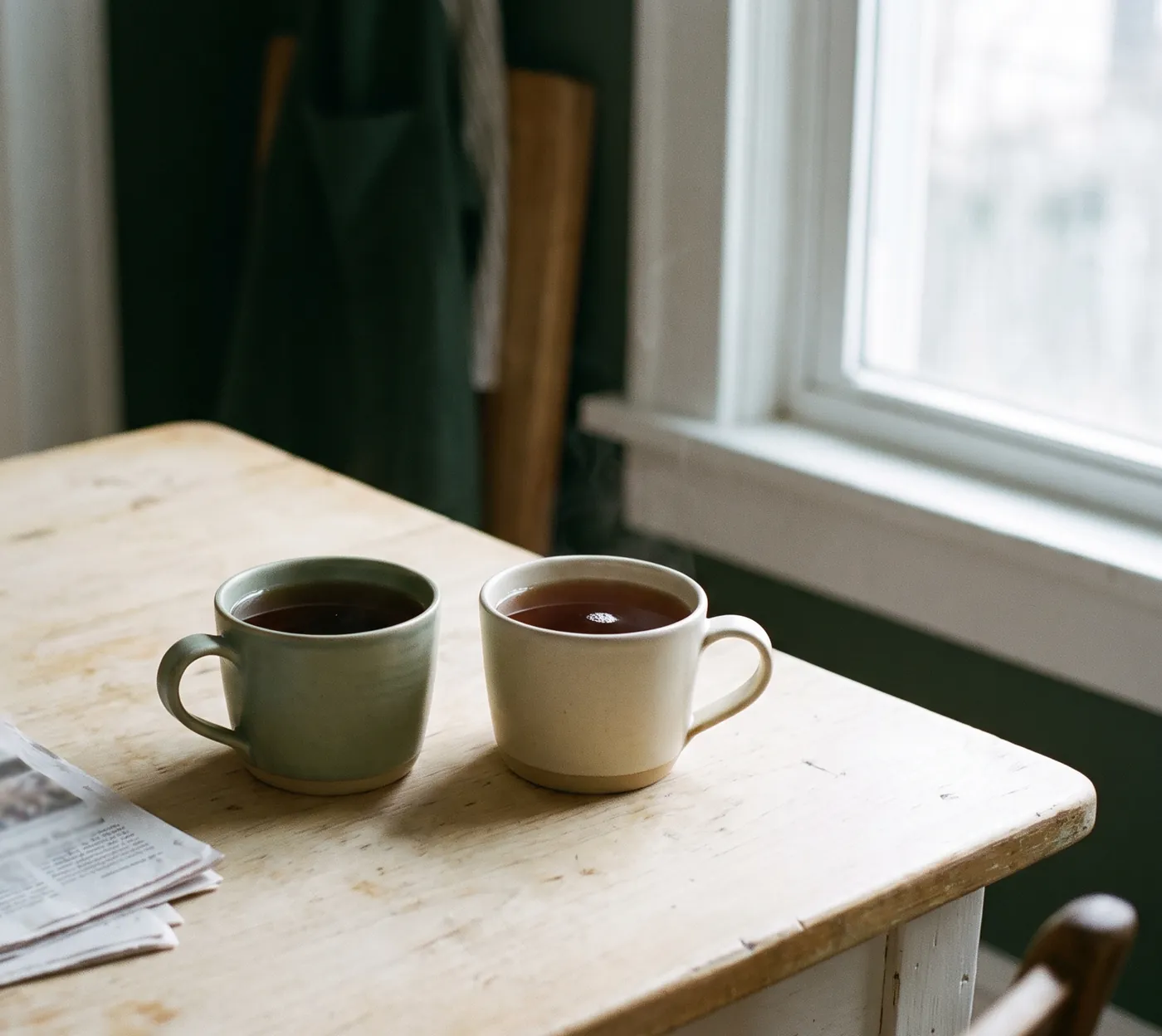 Two ceramic mugs of tea side by side on a pale wood kitchen table in soft morning light.